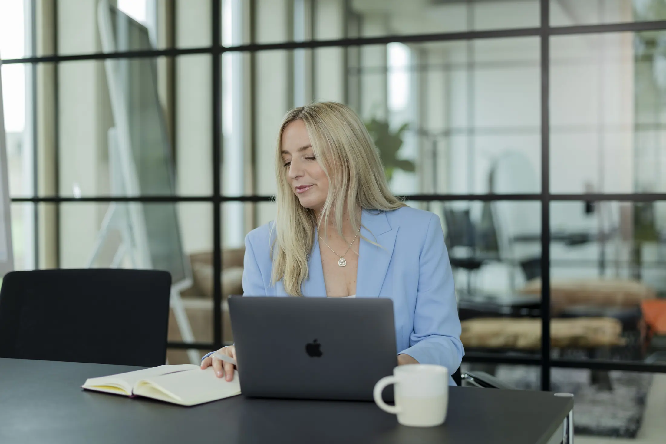 Frau im hellblauen Blazer arbeitet an einem Laptop, hinter ihr eine Glaswand mit Blick in den Büroraum