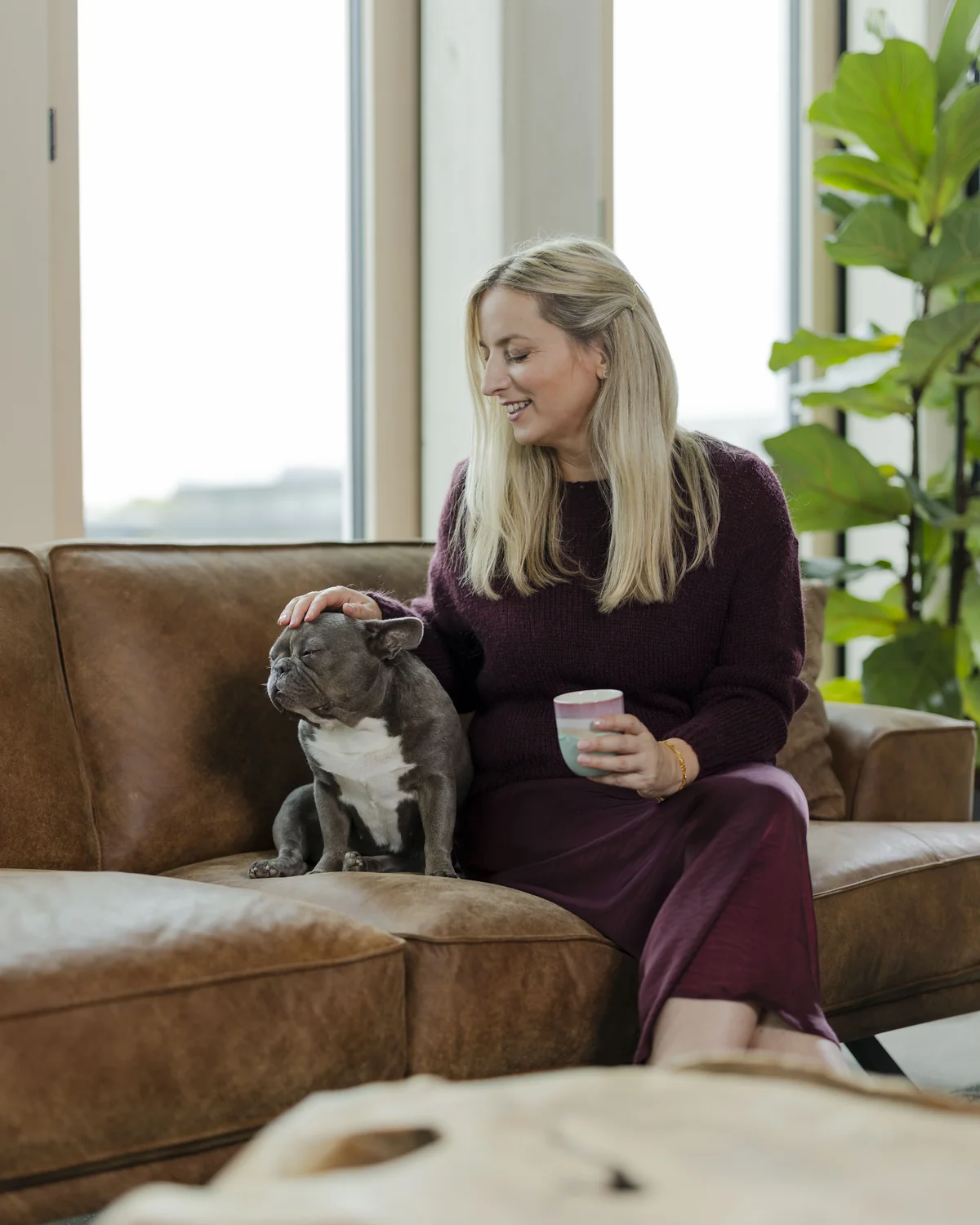 Frau im dunkelroten Kleid sitzt mit einer Tasse auf einem Ledersofa und streichelt einen kleinen Hund